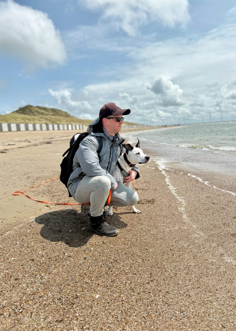 Hundeschule Partnerschnauze Mann kniet am Strand neben einem Hund und schaut aufs Wasser. Wolken am Himmel.