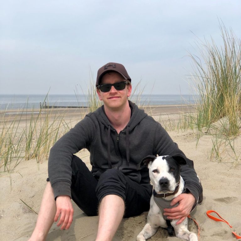 Hundetrainer Hauke Gehlen Mann sitzt am Strand mit seinem Hund, umgeben von Sand und Gras.